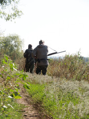 Ways to carry rifles. A hunters with a rifles walks along a country road. One of them has a rifle hanging on his shoulder, and the other one has it on the neck in front.