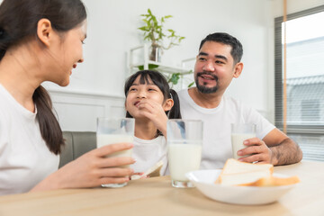 Asian  family enjoying breakfast at living room. little girl daughter sitting on table, drinking milk with smiling father and mother in morning. Happy family at home.