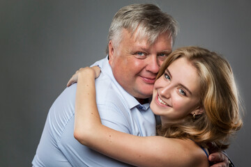 Dad and teenage daughter laugh and hug. A fat man in a shirt and a beautiful girl in a blue dress on a dark gray background. Love and tenderness. Close-up.