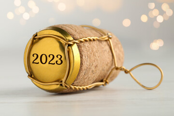 Cork of sparkling wine and muselet cap with engraving 2023 on table against blurred festive lights,...