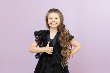 The little girl gives a thumbs up and smiles. A miss with wavy hair in a black dress on a pink...