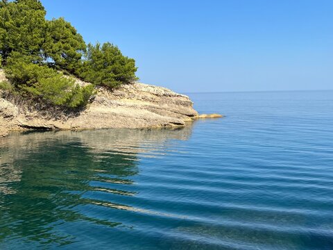 Sea ripples clear blue water near rocky island covered pine trees at sunny summer day.