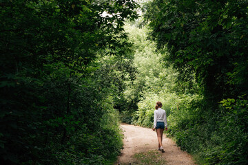 A young slender schoolgirl in denim shorts and a white shirt walks along a forest path into the distance, under an arch of trees.