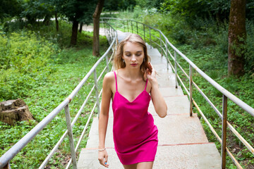 A slender young girl with long blond hair, in a pink dress, walks up the stairs, in the alley of the park, around green trees. He fixes his hair with his hand.