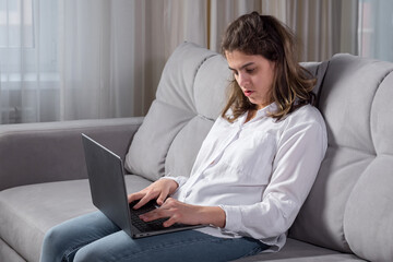 Young disabled woman with cerebral palsy works remotely on computer trying to type text by poorly mobile fingers sitting on sofa by window at home. - obrazy, fototapety, plakaty