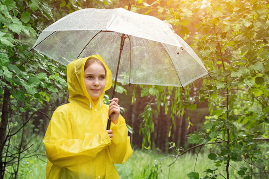 Cute girl in yellow raincoat holds umbrella to hide from rain. Happy teenager smiles enjoying rainy summer day in forest with lush greenery