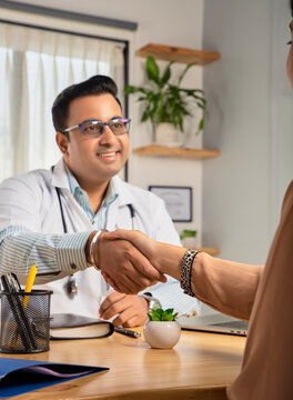 An Indian young man or male smiling physician or doctor sitting in a modern clinic wearing a stethoscope and apron shaking hand with a happy female patient. Medical, medicine, and healthcare concept