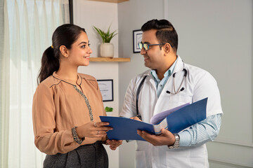 Asian Indian man or male physician or doctor wearing stethoscope and apron holding report file in hand and giving consultation to a smiling female or happy Woman patient. Medical, medicine, healthcare