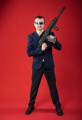 businessman with a gun. a guy in fashionable glasses, a suit holds a machine gun. Business concept. red background