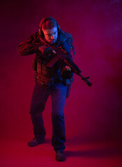 Fototapeta na wymiar soldier in full gear with weapons. a man in headphones, body armor, with a backpack and a belt. red background. colored, blue-red light