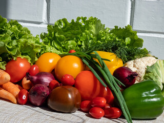 A pile of fresh, healthy and colorful vegetables in bright sunlight with white brick wall in background. Tomatoes, onions, carrots, peppers, cauliflower, salad, dill and greens