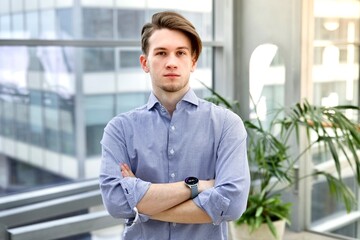 Portrait of handsome European young student or office worker employee in shirt standing indoors with his hands crossed looking at camera. University or college.