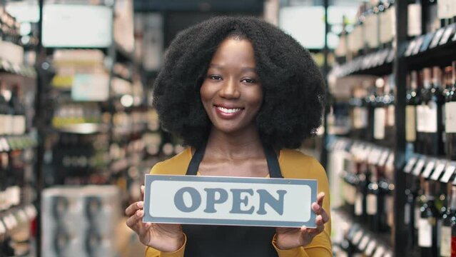 Portrait of african american female owner of wine store holding sign with word Open in her hands. Attractive smiling saleswoman welcoming customers at liquor market.