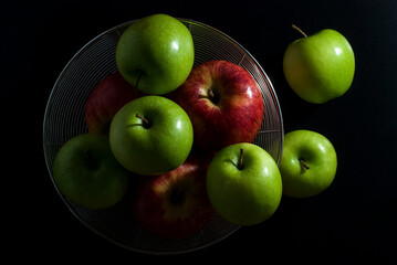 Red apples and green apples are placed in a stainless steel round basket and rest on a black background, top view.