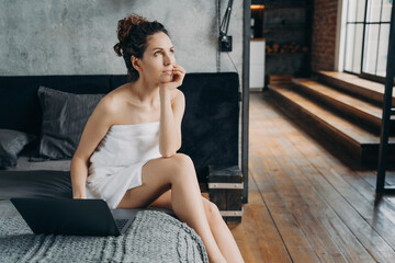 Caucasian girl wrapped in towel after bathing is sitting at laptop on her bed and dreaming.