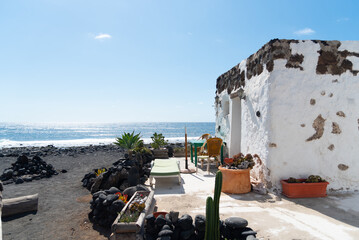 Casa tradicional de dintel blanco, situada en una playa de arena negra, típica de las Islas Canarias, Lanzarote en un día soleado con el mar al fondo. Paisaje volcánico y turismo de naturaleza. - obrazy, fototapety, plakaty