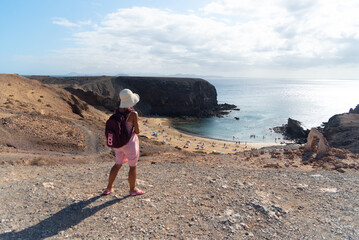 Una turista con gorro y pantalones cortos observando la playa de Papagayo en Lanzarote, Isla Canarias. Una mujer paseando por los acantilado y con la playa de fondo. 
