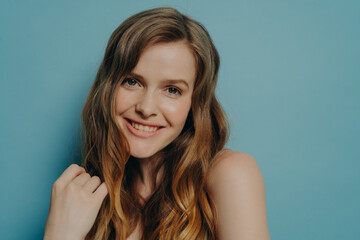 Fototapeta premium Studio portrait of cute teen girl with wavy hair smiling and looking at camera, isolated on blue
