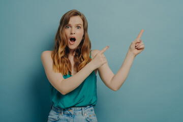 Fototapeta premium Surprised young woman in casual clothes pointing to right side with index fingers, isolated on blue