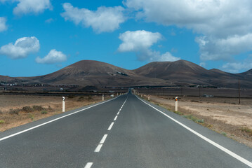 Carretera de dos carriles, recta, cruzando un impresionante paisaje desértico y volcánico, al fondo una gran montaña volcánica durante un día soleado con el cielo azul en Lanzarote, Islas Canarias