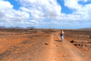 Fototapeta na wymiar Mujer rubia de espaldas que lleva una mochila atravesando el paisaje volcánico y desértico de Lanzarote con volcanes inactivos al fondo durante un día soleado en las Islas Canarias.