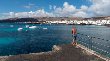 Mujer rubia pescando frente a Punta Mujeres  con peque&ntilde;as casas blancas t&iacute;picas de Lanzarote en la costa junto al mar turquesa con barcos y volcanes inactivos detr&aacute;s un d&iacute;a soleado en Islas Canarias