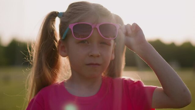 Upset little girl takes off sunglasses to show injured eye at rural site closeup slow motion. Unfriendly bug bite. Summer sunset time