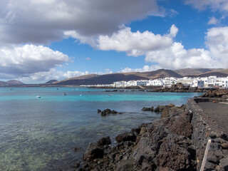 Fototapeta na wymiar Pueblo de Punta Mujeres con casas tradicionales de color blanco en la isla de Lanzarote en Islas Canarias rodeado por un mar turquesa y volcanes inactivos al fondo durante un día soleado y despejado.