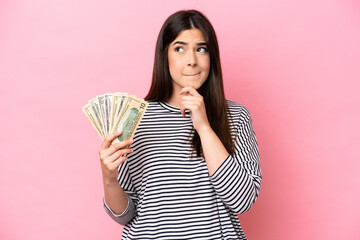 Young Brazilian woman taking a lot of money isolated on pink background having doubts and thinking