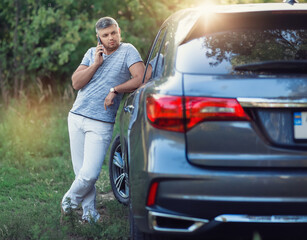 A middle-aged man is standing near the car. Muccina speaks on the phone. Dear beautiful car. The man is calm and confident. The car is in the summer park.