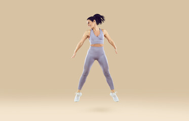 Fototapeta na wymiar Woman in sporty short top and gym leggings doing sports exercise and jumps up on beige background. Full length of smiling brunette woman doing sport jumping isolated on colored background. Banner.