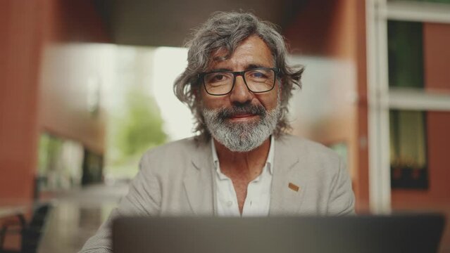 Mature businessman with beard in eyeglasses sits in cafe. Middle aged manager successful man working on laptop pc computer sit at cafe outdoor. Raises his eyes and smiles at the camera