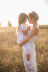 A cute little girl with long blond curly hair and her mother in a white summer dress and a straw boater hat in a field in the countryside in summer at sunset. Nature and Ecolife