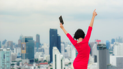 Fototapeta na wymiar Back side of a short-haired business women wearing a red dress, holding a tablet, jumping joyfully, dancing on a rooftop with blur background of high-rise buildings.