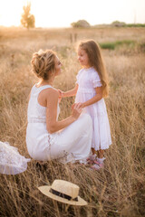 A cute little girl with long blond curly hair and her mother in a white summer dress and a straw boater hat in a field in the countryside in summer at sunset. Nature and Ecolife