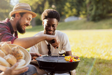 A bearded middle-aged guy is preparing a barbecue for friends. A group of college guys spend time together surrounded by nature.