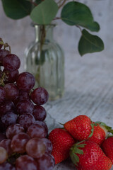 Appetizing, fresh, juicy, ripe grape and strawberry assortment on light gray background. Glass vase with greenery in the background.