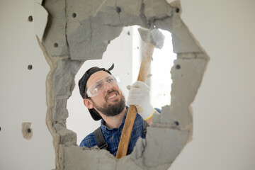 A smiling guy demolishes a wall in a house using a construction hammer on a long stick. Fun demolition of an apartment during renovation, getting rid of an unnecessary wall gaining space.