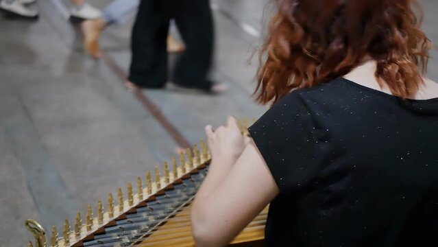 Young red-haired female street musician plays a melody on a plucked string instrument gusli or zither while sitting on the street and putting the musical instrument on her knees