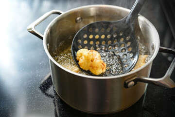 Cauliflower floret on a spatula deep fried in a pot with steaming cooking oil on the stove,...