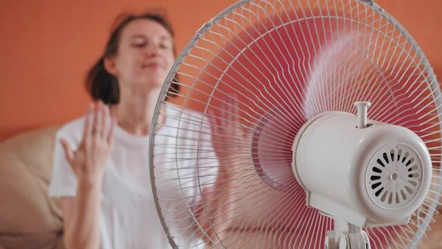 Slow motion, young caucasian woman in white t-shirt sits in front of electric home propeller cooler fan, waving hands, air stream blowing short hair, lady getting cool down and relax in hot apartment.