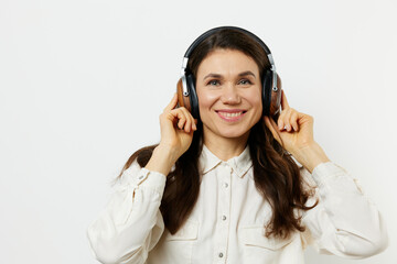an emotional, beautiful, pleasant woman stands in a white shirt listening to music and holding headphones with her hands, smiling pleasantly with pleasure.