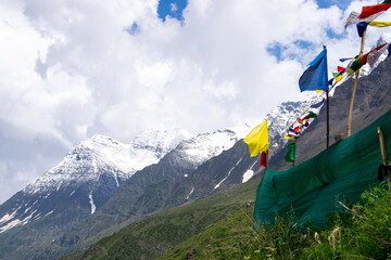 Beautiful multi colored prayer flags are flying through strong winds in the higher range of himalayas. Snow capped mountains peaks all covered in dense clouds and a little of blue skies can be seen.
