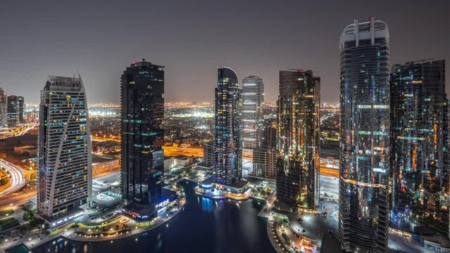 Tall residential buildings panorama at JLT district aerial night timelapse, part of the Dubai multi commodities centre mixed-use district. Illuminated skyscrapers around pond