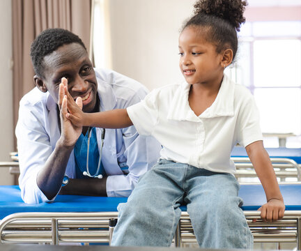 Child patient sitting on hospital bed with African doctor for medical care. Smiling little girl happy healthy after professional checkup at clinic. Practitioner, pediatrician checking, examining kid