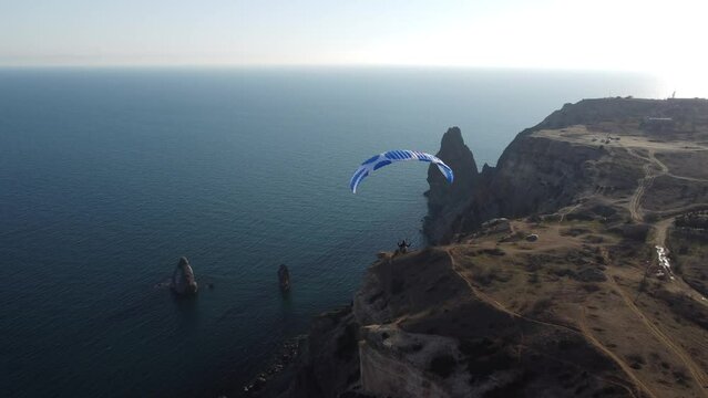 Aerial drone view of a man flying a white and blue paraglider over a hill and trees to the sea waves near the rocks. Active paraglider flight over the seascape with clear skies at suset. Extreme sport