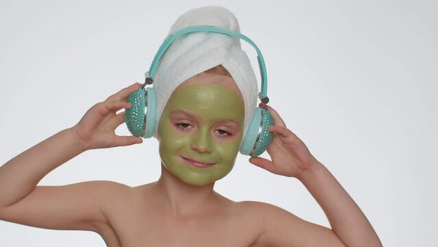 Smiling young child girl after bath in towel on head and moisturizing green mask on face listening to music on headphones, dancing. Teenager kid skin care treatment, natural cosmetics. Female portrait