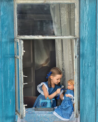 Portrait of girl kid tailor sew making doll's clothes on a children's sewing machine in the window of an old wooden house.