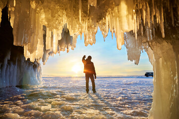 View from the ice cave with huge icicles on the frozen Lake Baikal at sunset. A man stands with his back to the cave and admires nature. Winter travel or freedom concept. 