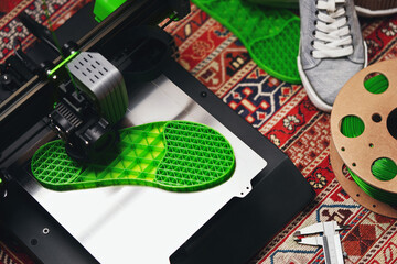 A shoe sole with distinct inner structure is printed from green filament on a 3D-printer with steel print bed. comfy home concept with persian rug, grey sneaker shoes and filament. selective focus - obrazy, fototapety, plakaty